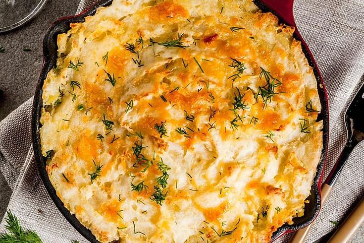 a casserole with cheese and herbs in a red dish on a cloth next to utensils