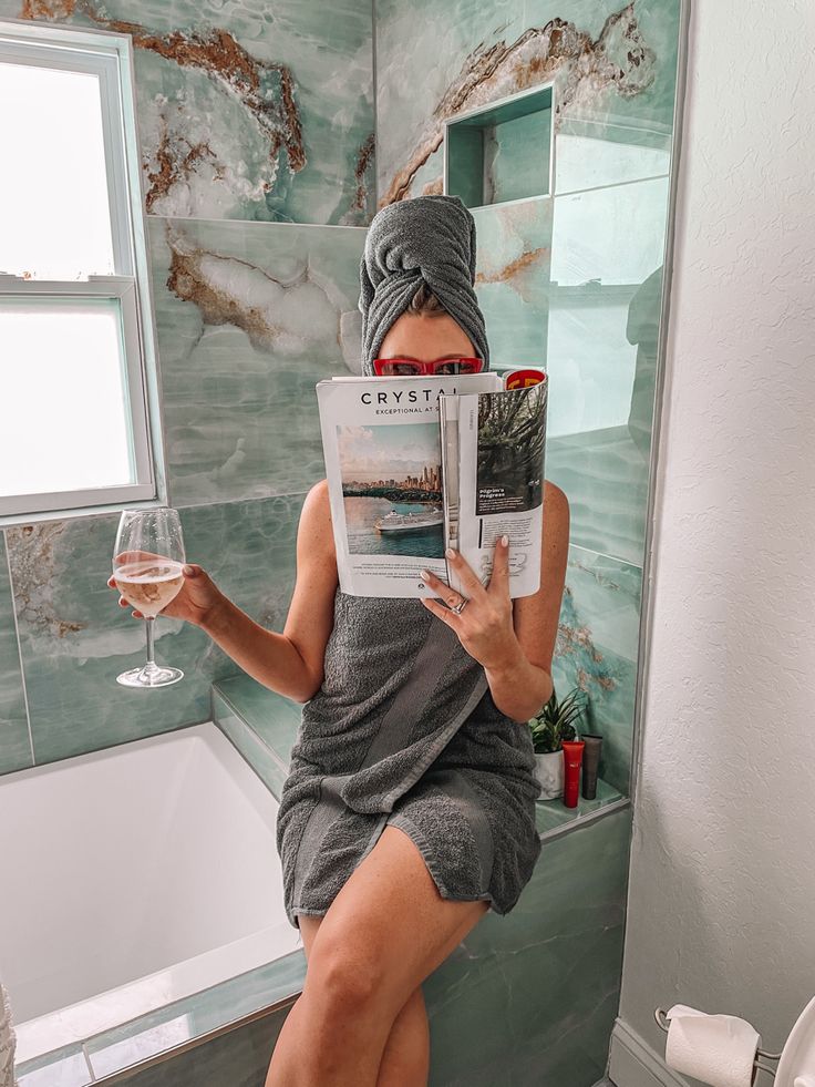 a woman sitting on a toilet reading a magazine and holding a glass of wine in her hand