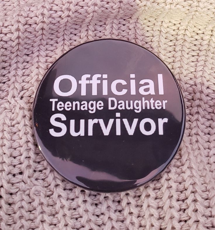 a black button with the words official teenage daughter survivor on it sitting on a knit surface