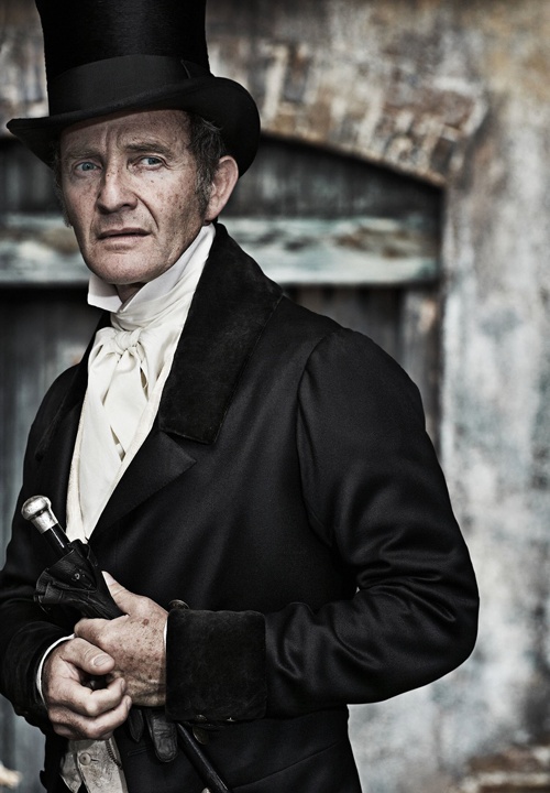 an old man wearing a top hat and holding a pipe in his right hand while standing next to a building