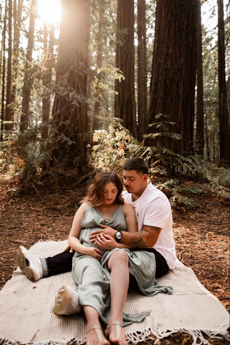 a man and woman are sitting on a blanket in the woods with sun shining through the trees