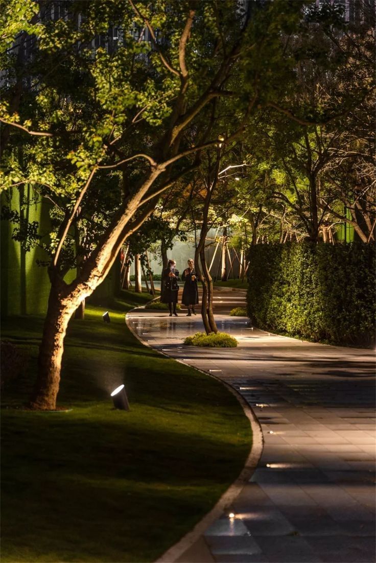 two people walking down the street at night with trees and grass on either side of the road