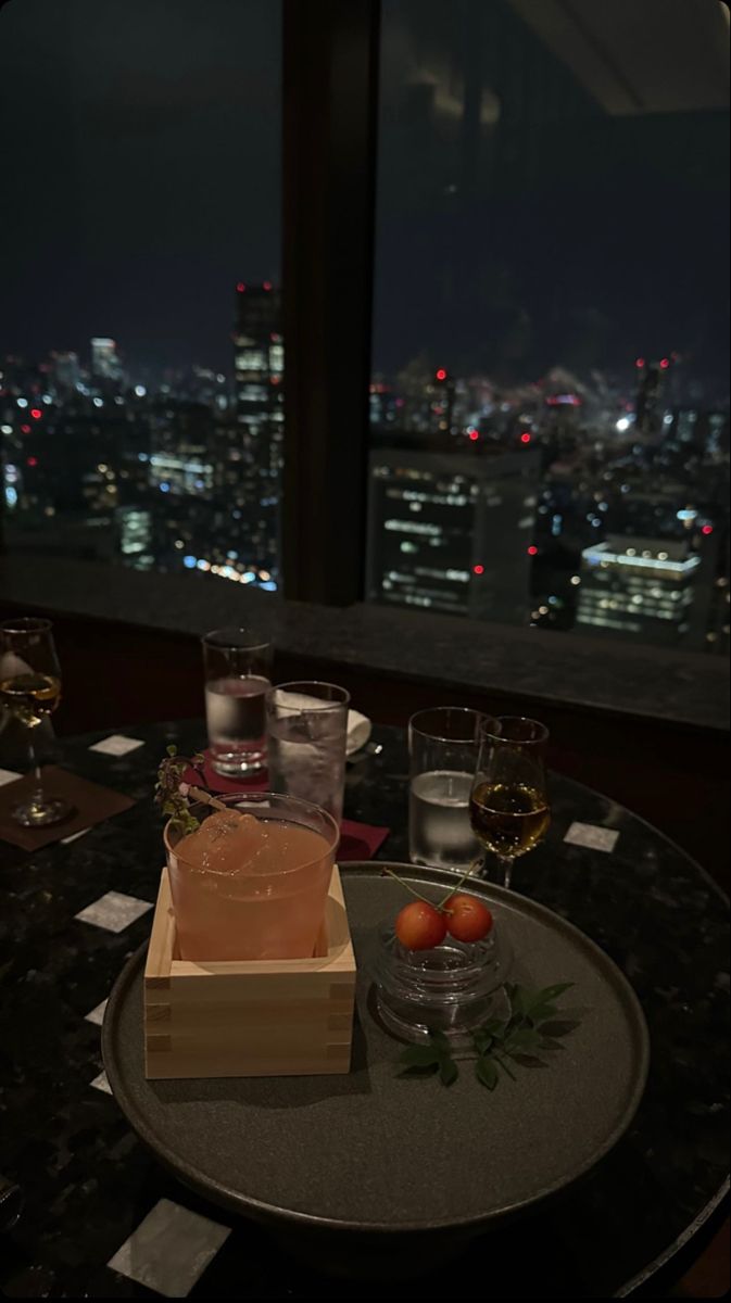 a table with drinks and fruit on it in front of a window overlooking the city at night