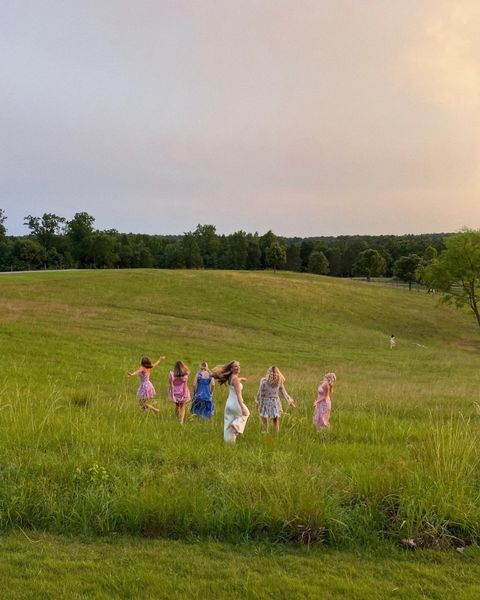 four girls are walking in the grass with their arms around each other and one girl is flying a kite