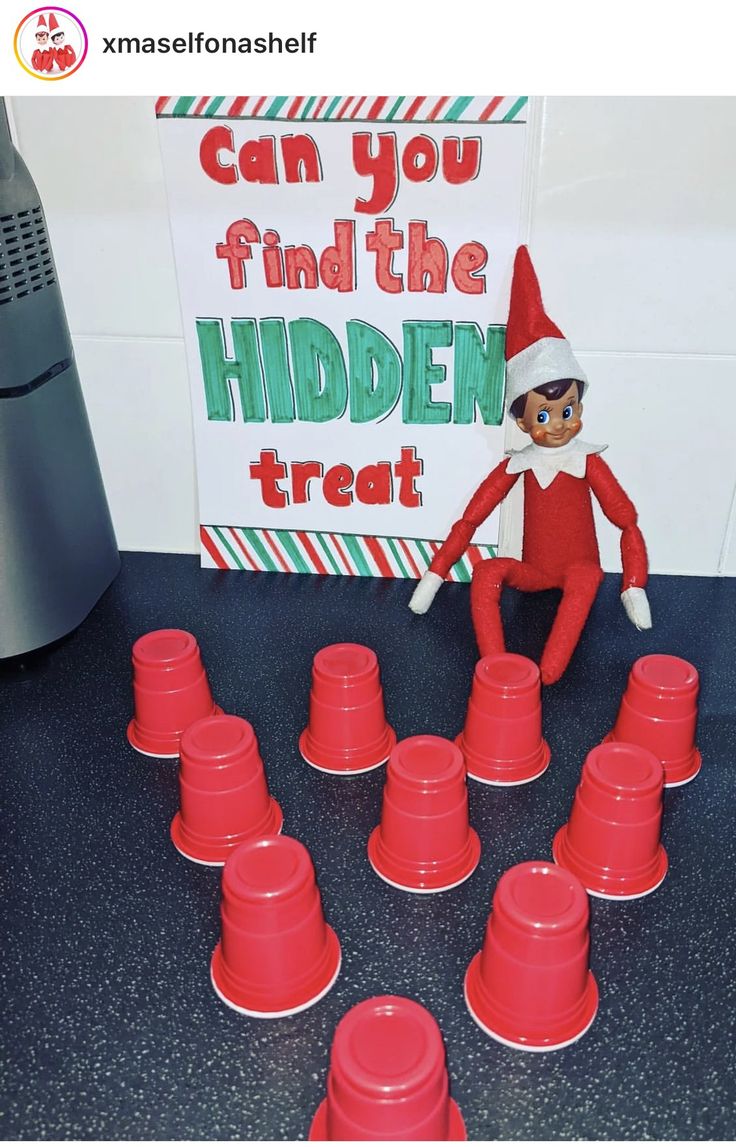 an elf is sitting on top of red cups and has a sign that says can you find the hidden treat?