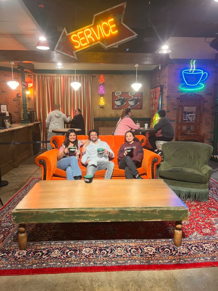 three people sitting on an orange couch in front of a coffee table with a neon sign above it