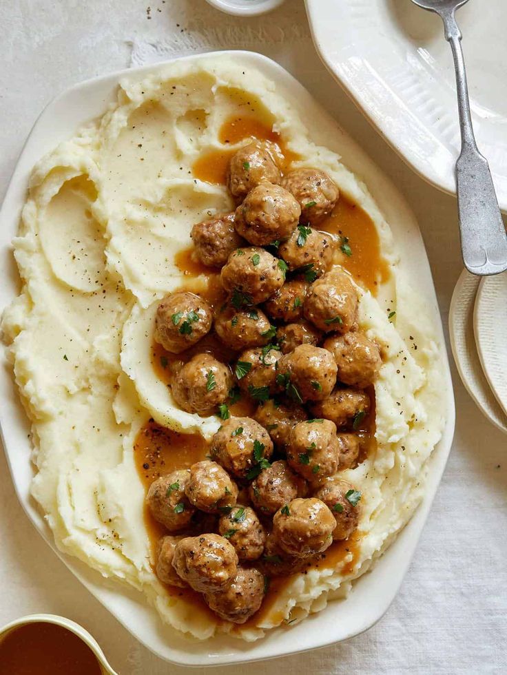 mashed potatoes with meatballs and gravy on a white plate next to silverware