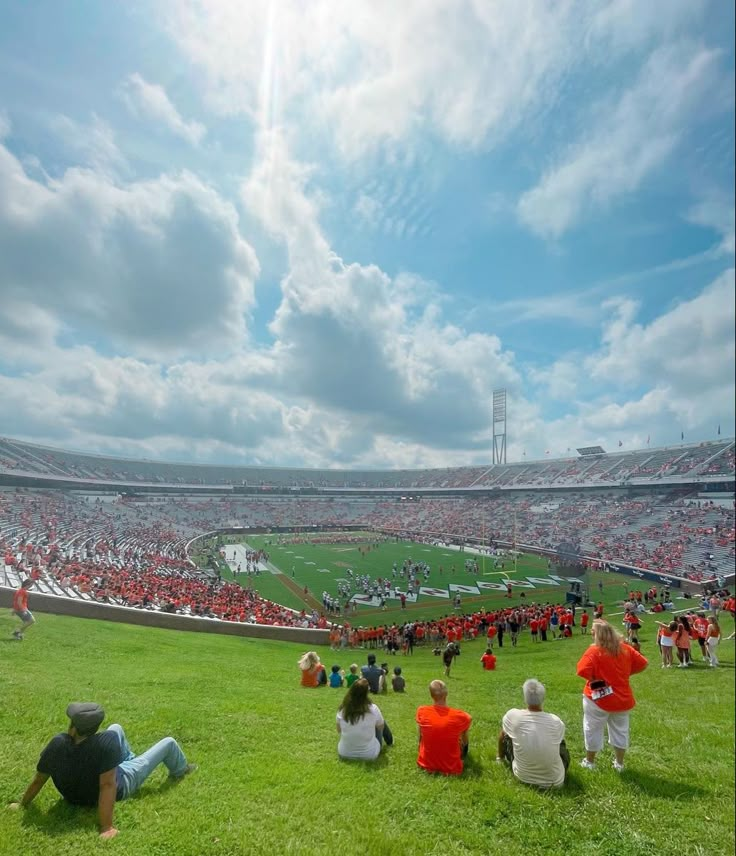 many people are sitting on the grass in front of an empty stadium with red seats