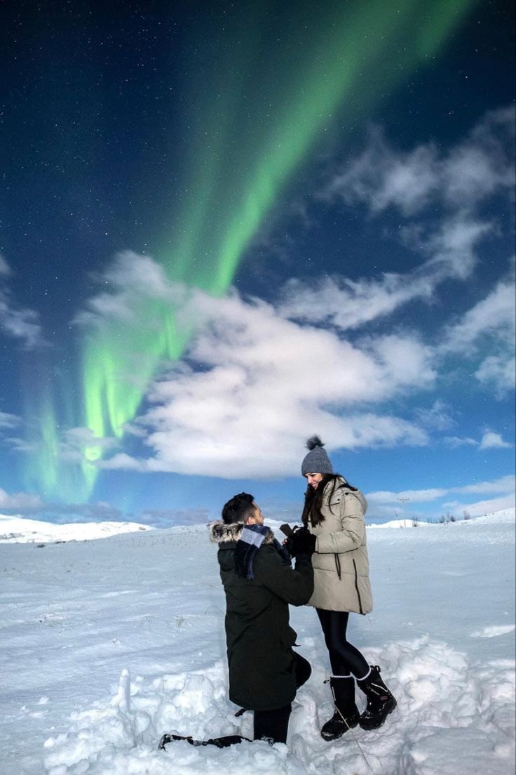 two people are standing in the snow with an aurora bore above them and looking at something