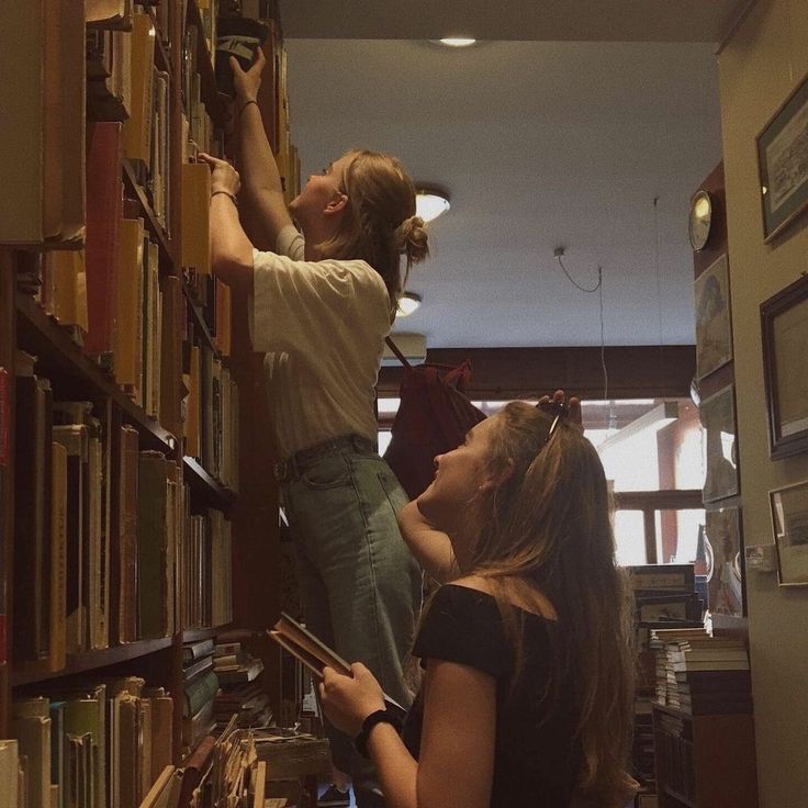 two girls are looking at books on the shelves in a library and one girl is reaching for them