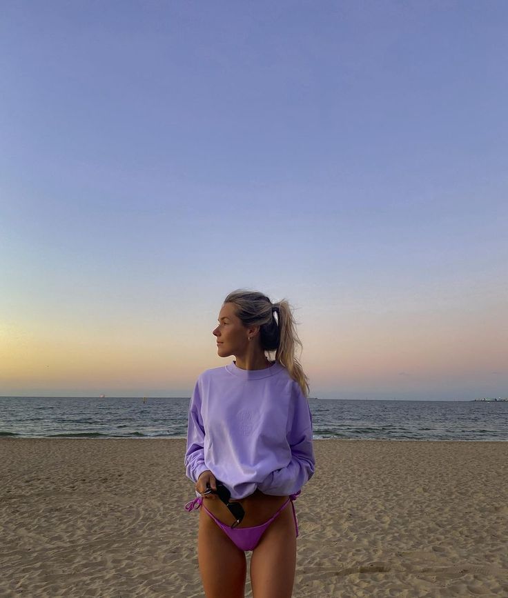 a woman standing on top of a sandy beach next to the ocean with her hands in her pockets