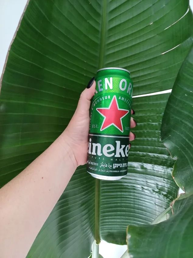 a person holding up a can of beer in front of a large leafy plant