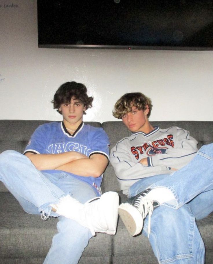 two young men sitting on top of a gray couch in front of a flat screen tv