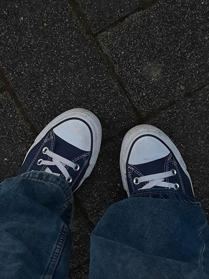 a person wearing blue jeans and white sneakers standing on the pavement with their feet crossed