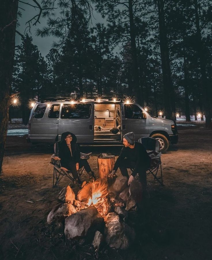 two people sitting around a campfire in front of a van at night with lights on