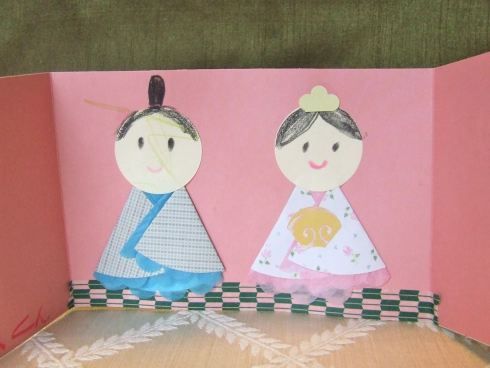 two paper dolls sitting on top of a wooden table next to a pink box with white trim