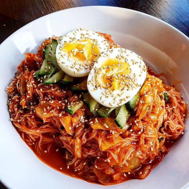 two eggs on top of noodles in a white bowl with red sauce and sesame seeds