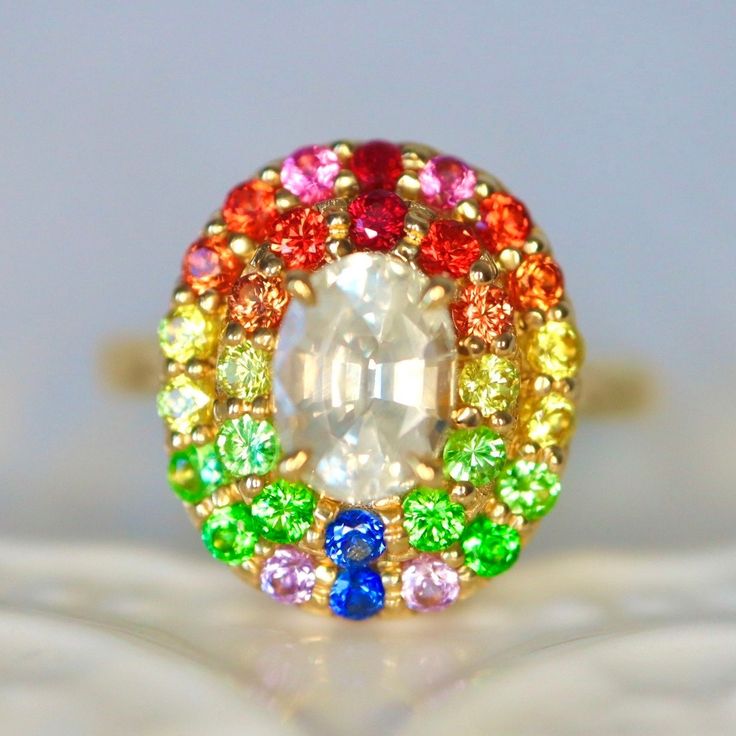 Opalescent White Sapphire and rainbow color halo ring Condition: Brand New & Custom Designed by me Size: US 7 sizable Metal: Solid 14K Yellow Gold Center Stone: Natural White Sapphire. Opalescent white color. Internally flawless.  Side Stones: Red rubies, orange sapphires, neon orange sapphires, yellow sapphires, mint garnets, tsavorite garnet, blue sapphires, purple sapphires 1.5ct in total. Measurement: 15*13mm face. 7mm height. Band Width: 2.4mm on the back Band Thickness: 1.4mm on the back C Multicolor Oval Heirloom Ring, Heirloom Multicolor Oval Rings, Heirloom Style Multicolor Oval Rings, Multicolor Round Jewelry With Halo Setting, Multicolor Jewelry With Halo Setting, Multicolor Oval Halo Setting Jewelry, Oval Multicolor Jewelry With Halo Setting, Rainbow Oval Multi-stone Jewelry, Multicolor Oval Jewelry With Halo Setting