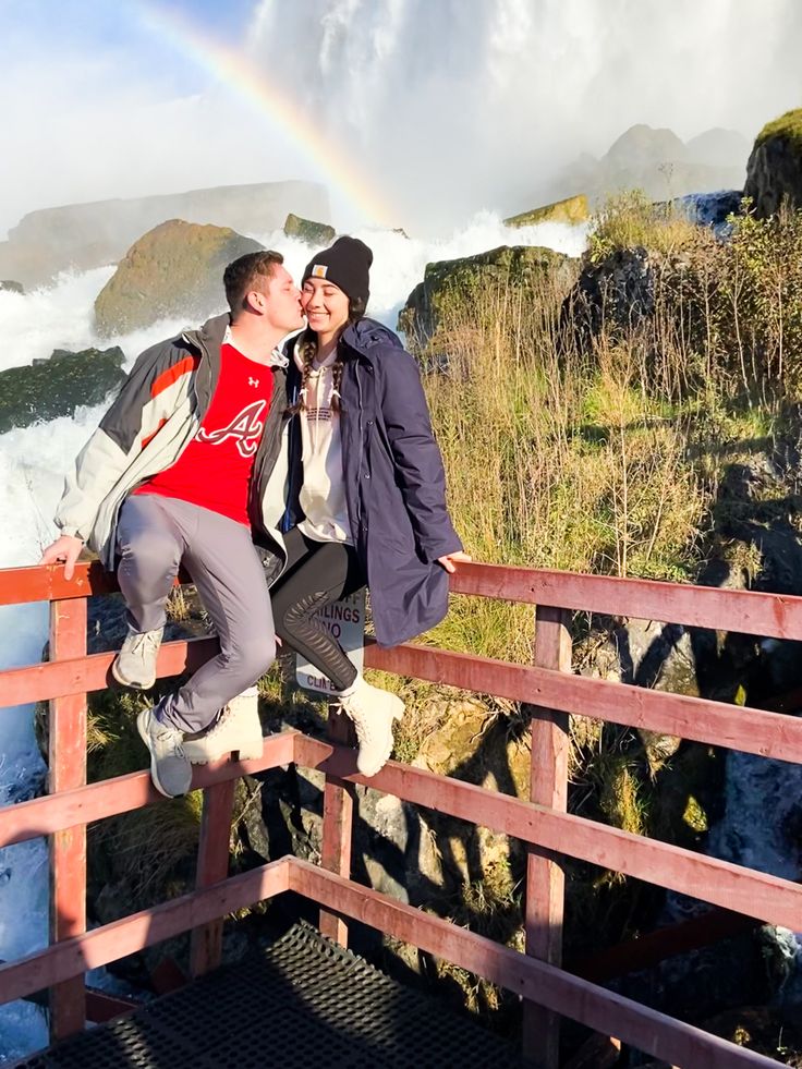 two people are standing on a bridge with a rainbow in the background and water crashing behind them