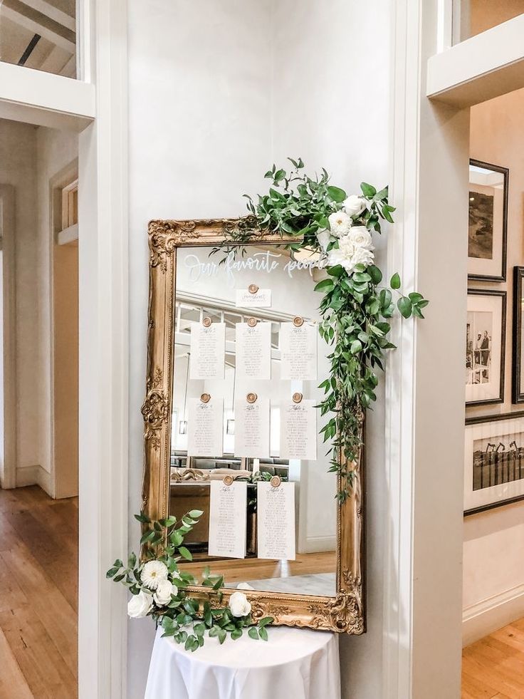 a mirror hanging on the wall next to a table with white flowers and greenery