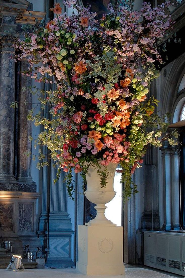 a tall white vase filled with lots of flowers on top of a floor next to a wall