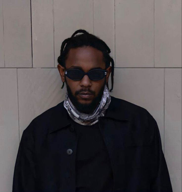 a man with dreadlocks wearing sunglasses and a scarf around his neck standing against a wall