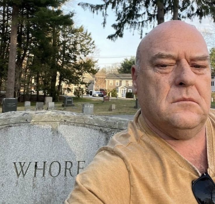 a bald man with a tie standing in front of a headstone