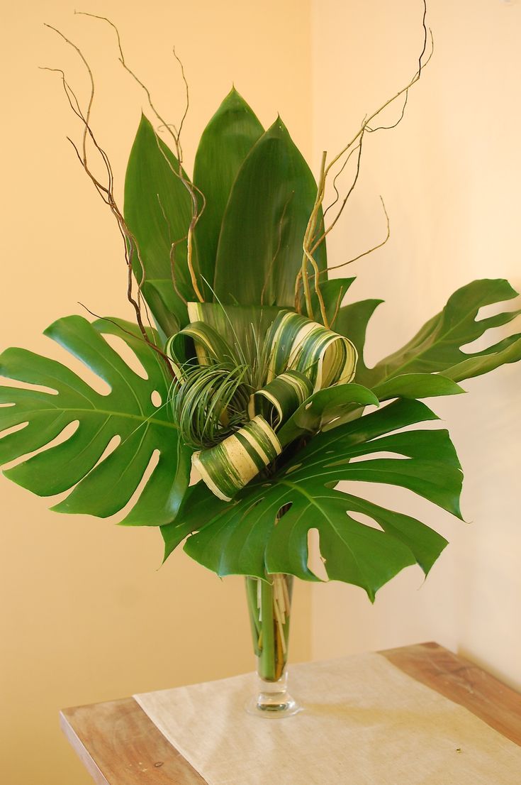 a vase filled with lots of green leaves on top of a wooden table next to a wall