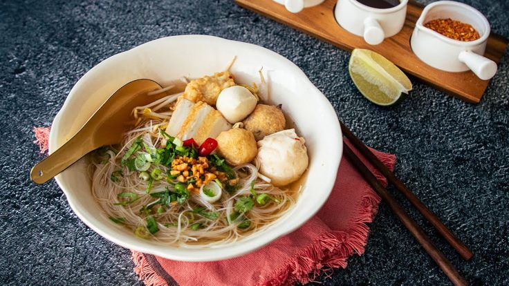 a white bowl filled with noodles, meat and veggies next to chopsticks