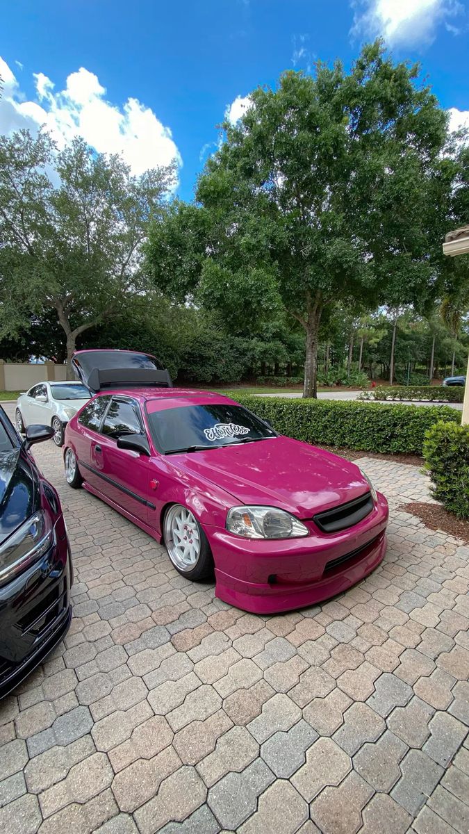 two pink cars parked next to each other on a brick driveway with trees in the background