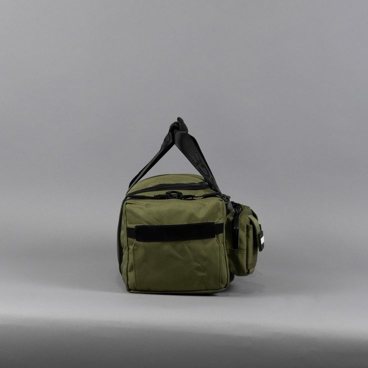 an olive green duffle bag with black straps on the front and side pockets, sitting against a gray background