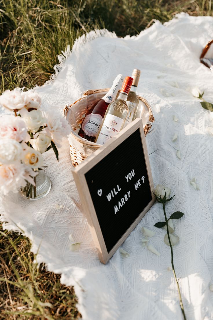 a picnic blanket with bottles of wine and flowers on it, next to a sign that says will you marry me?