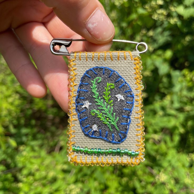 a hand is holding a small piece of beaded fabric with an image of a plant on it