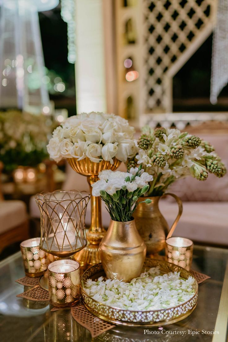 a table topped with gold vases filled with flowers and white flowers next to candles