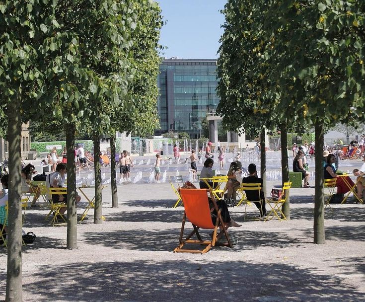 many people are sitting and standing around in the park with umbrellas, chairs, and trees