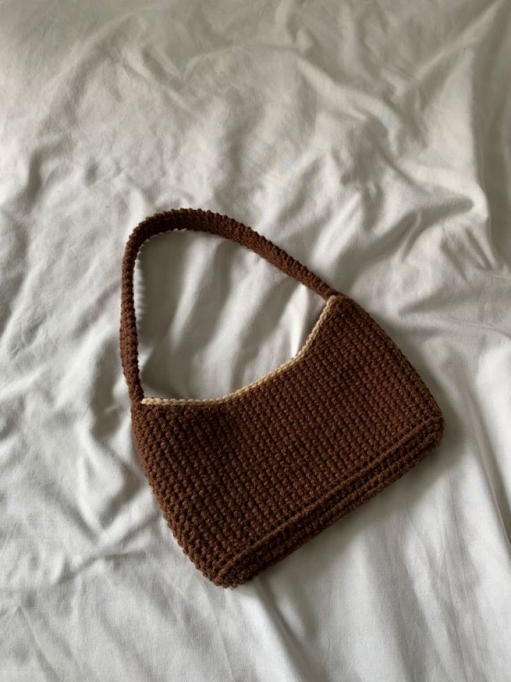 a brown crocheted purse laying on top of a white bed with sheets and pillows