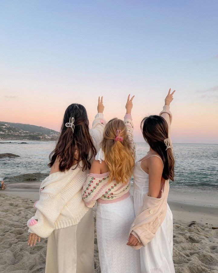 three girls standing on the beach with their arms in the air and one girl wearing a white dress