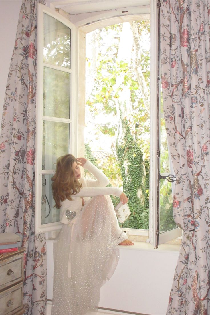 a woman standing in front of a window next to a white dresser and curtained windowsill