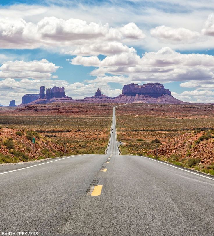 an empty road in the desert with mountains in the background and clouds in the sky