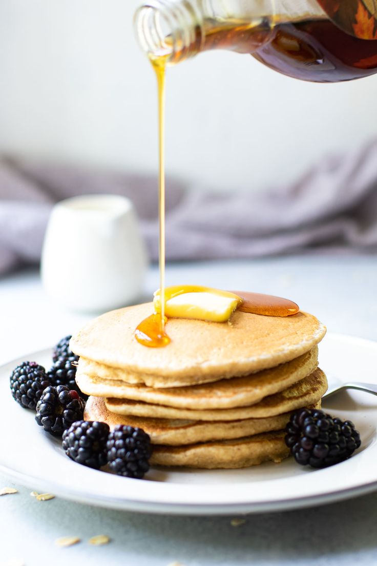 a stack of pancakes with syrup being drizzled over them and blackberries on the side