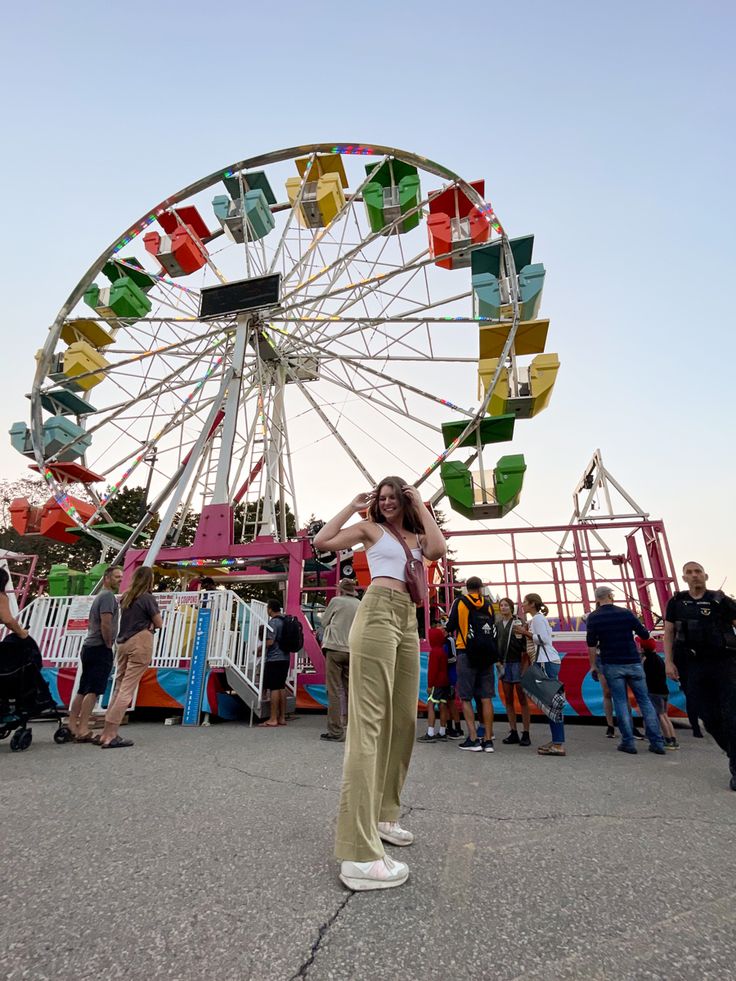 a woman standing in front of a ferris wheel at an amusement park with people walking around