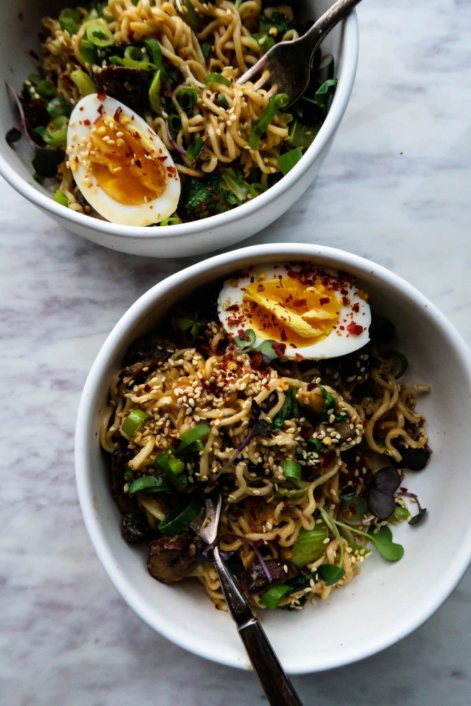 two bowls filled with noodles and vegetables on top of a marble countertop next to an open hard boiled egg