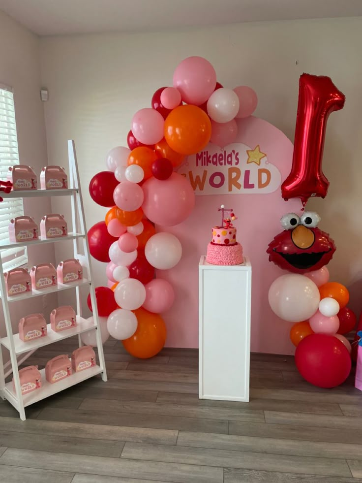 the balloon arch is decorated with balloons and pink, orange, and red items for a first birthday party