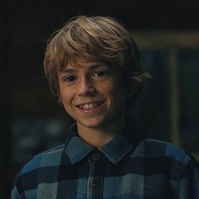 a young boy with blonde hair smiles at the camera while wearing a blue and black checkered shirt