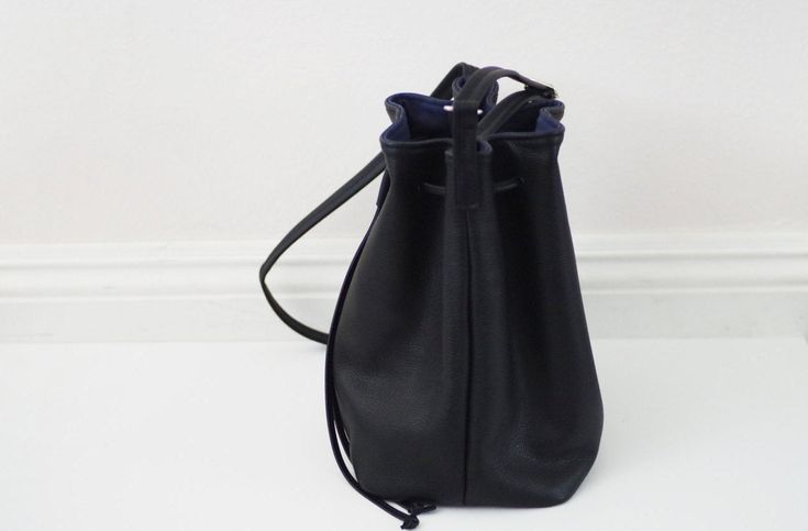 "This bucket bag features soft black leather, cotton lining and a drawstring closure. Carry your phone, a wallet, keys and small items in this bag. Details: -Dimensions: 10.5\" length (top to bottom), 13.5\" opening top width, 6\" depth, 7.5\" X 6\" base (bottom) -Shoulder strap measures approx. 45-50\" total (shoulder strap is adjustable) -Soft black leather -Cotton lining -One internal pocket Hardware color: -Silver (pictured) -Gold Options: -Real cowhide leather (pictured) -Vegan (fake) leath Casual Leather Backpack With Phone Bag, Casual Leather Backpack With Phone Bag For Everyday, Black Pouch Bucket Bag For Everyday Use, Black Leather Bucket Bag With Leather Handles, Black Leather-handled Bucket Bag, Leather Drawstring Bag With Removable Pouch For Travel, Leather Drawstring Bag For Travel, Soft Leather Drawstring Travel Bag, Soft Leather Drawstring Bag For Travel