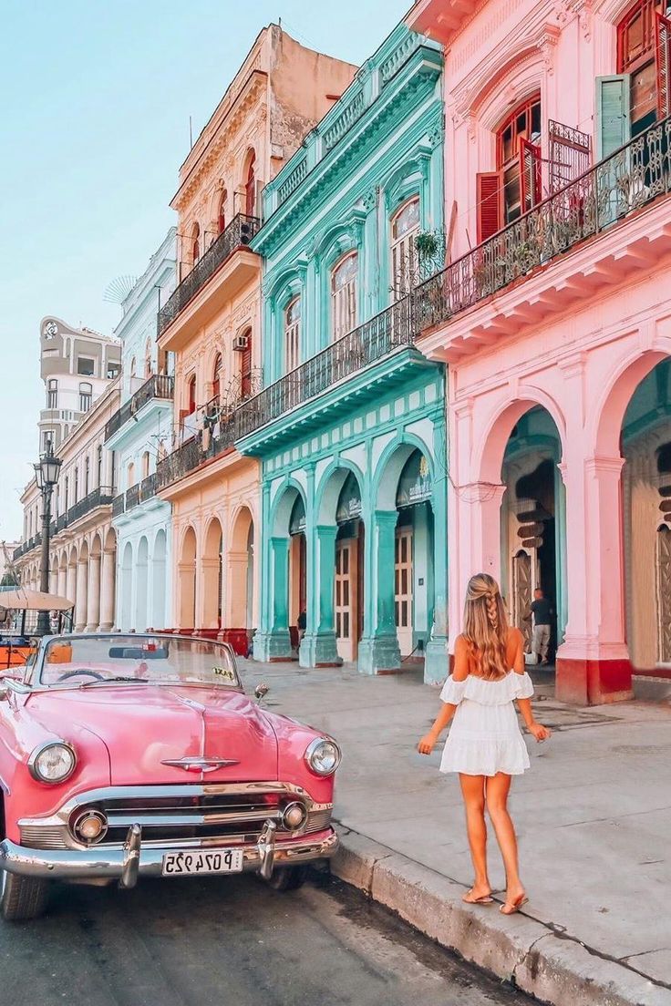 a woman standing next to an old car on the side of the road in front of colorful buildings