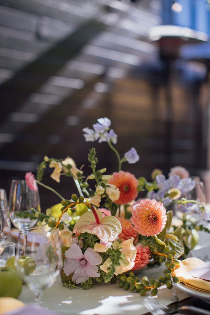 an arrangement of flowers and greenery on a table with wine glasses, plates and utensils