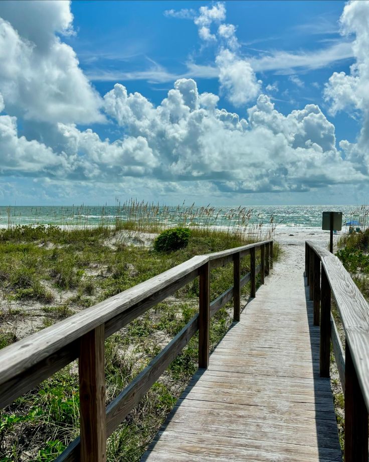 a wooden walkway leading to the beach with clouds in the sky and grass on either side