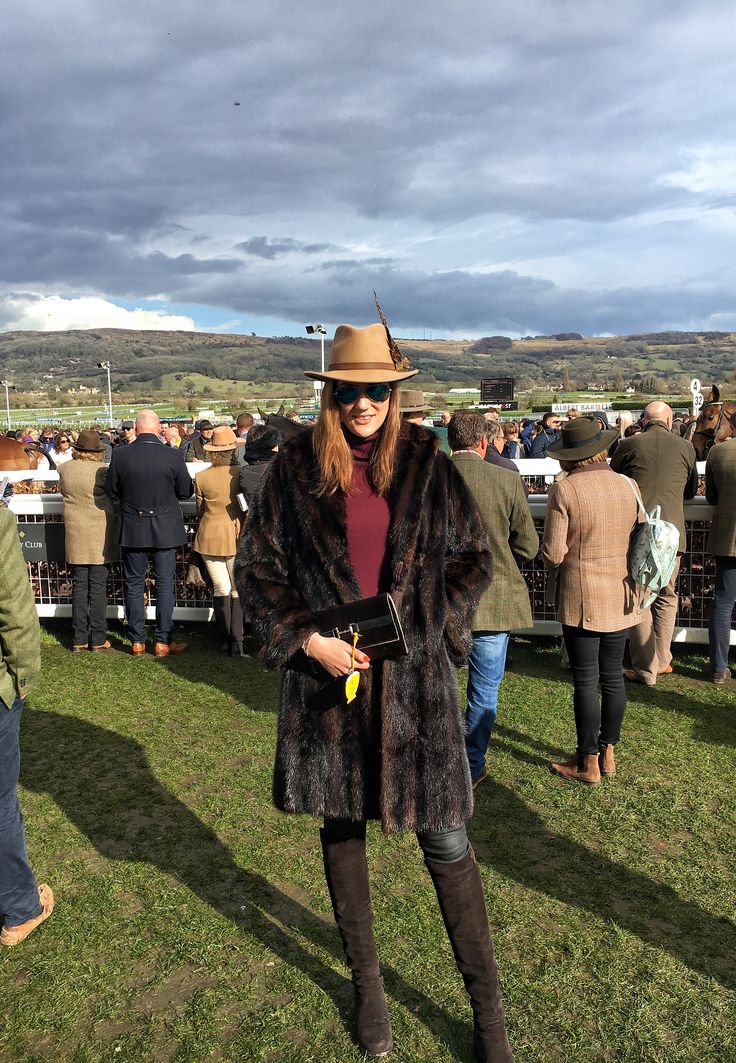 a woman in a fur coat and hat standing on grass with other people behind her