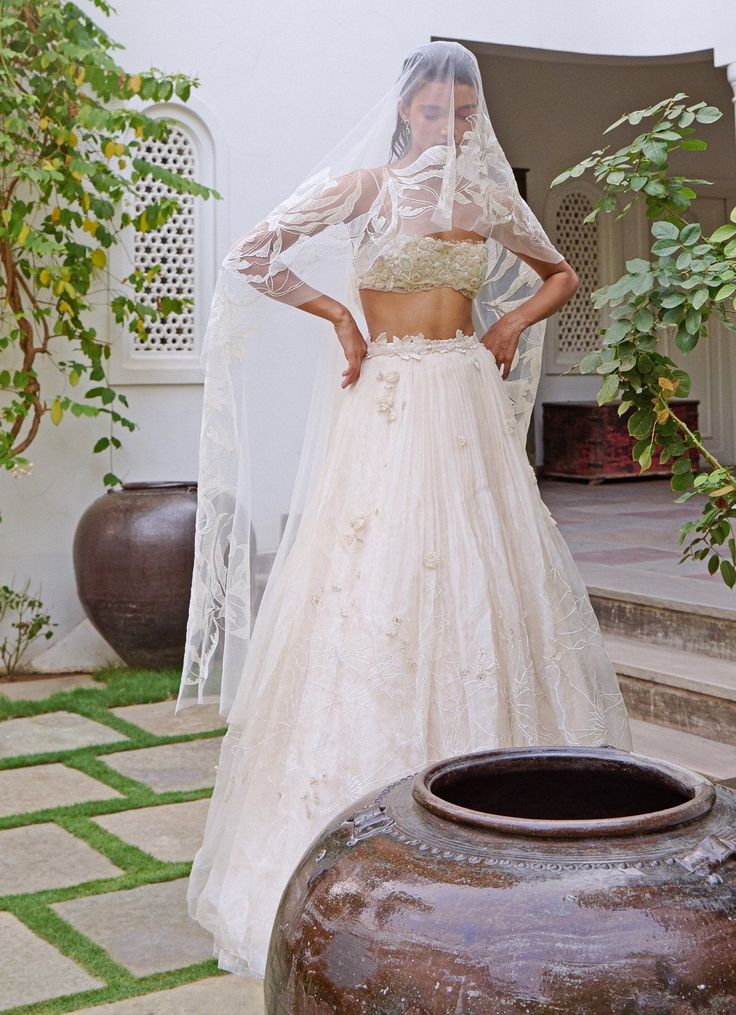 Elevate your style with the stunning Ivory Floral Embroidered Lehenga Set, a breathtaking blend of traditional elegance and modern sophistication. Crafted from luxurious organza, this ivory lehenga is layered with soft silk and French lace, creating a rich, textured base. Intricate tone-on-tone threadwork and zardosi embroidery enhance the fabric, while hundreds of shimmering, hand-cut organza flowers are scattered throughout, adding a whimsical, ethereal charm. The waistline, adorned with delicate floss embroidery and hand-appliquéd beads, exudes opulence and grace. Paired with a strapless crop blouse, featuring sparkling beads and intricately hand-cut organza flowers. With peek-a-boo lace details and an underlayer of fine French lace and silk, the blouse is both romantic and contemporary Ivory Lehenga, Zardosi Embroidery, Organza Flowers, Embroidered Lehenga, Indian Wedding Wear, Indian Outfit, French Lace, Crop Blouse, Peek A Boo